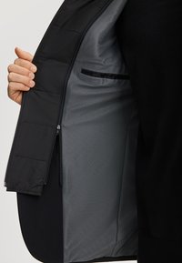 Czarna kurtka z pikowaną podszewką, posiadająca szaro-sieciowy środek i kieszeń na zamek. Materiał jest gładki o matowym wykończeniu.