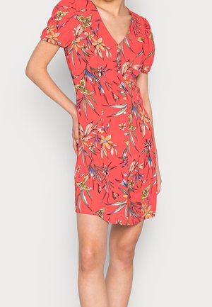 Robe rouge à manches courtes avec col en V et imprimé floral, portée par une personne avec une main sur la hanche.