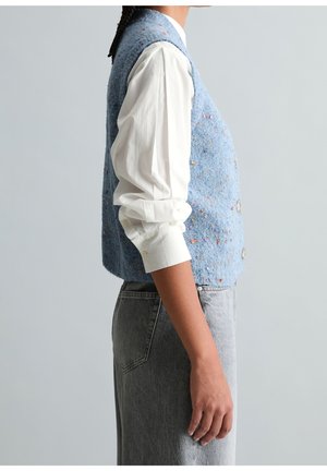 Personne portant une chemise blanche à manches longues, un gilet texturé bleu clair boutonné et un jean gris taille haute, montré de profil.