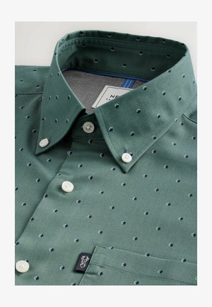 Chemise verte à manches courtes en tissu texturé, présentant un motif à pois subtils en bleu marine, avec des boutons blancs et une poche à rabat.