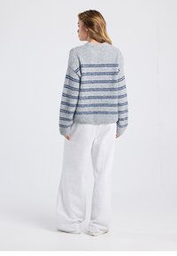 Grauer Strickpullover mit marineblauen Streifen, Rundhalsausschnitt und lockerem Sitz, kombiniert mit locker sitzenden weißen Jogginghosen. Weiche Textur und gerippte Bündchen.