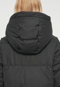 Čierna prešívaná bunda s vysokým golierom a nadrozmernou kapucňou, ktorá má hladkú, matnú textúru a prešívané detaily na chrbte.