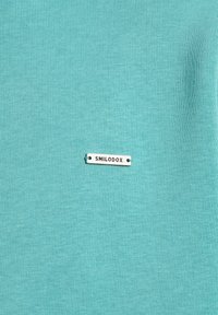 Un tissu turquoise clair avec une texture lisse et un subtil relief, comportant une étiquette argentée portant la mention "SMILODOX".