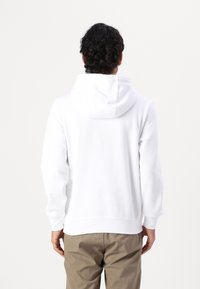 Felpa bianca in cotone con cappuccio con coulisse, tessuto morbido, polsini e orlo a coste, indossata sopra pantaloni beige, vista da dietro.