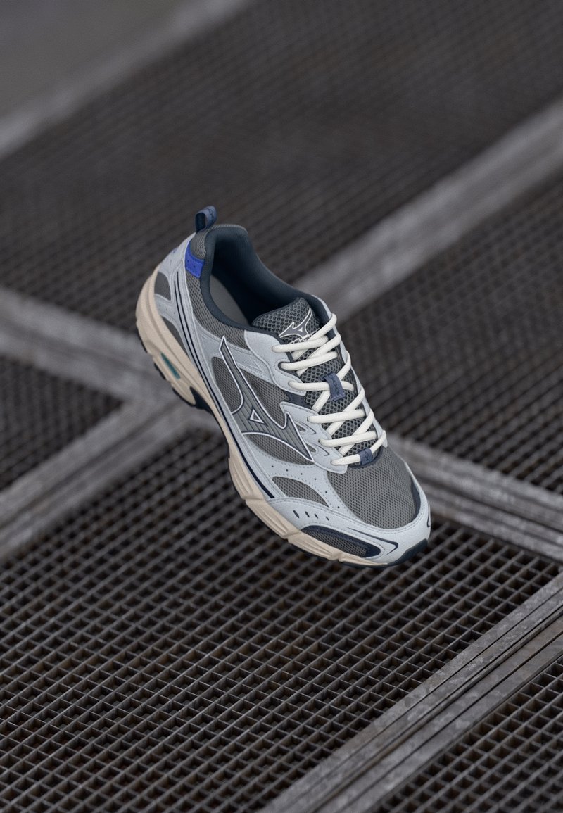 Grey running shoe with mesh panels, white laces, and a blue accent on the heel. Features a cushioned sole and structured design details.