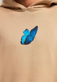 Beige hættebluse med et blåt sommerfugl grafik på brystet. Stoffet ser blødt ud med en tekstureret finish og en hætte med snøre.