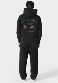 Persona con sudadera negra que lleva la inscripción "LA COURSE" y gráficos de un caballo alado, combinada con pantalones negros y zapatillas blancas, de pie mirando hacia atrás.