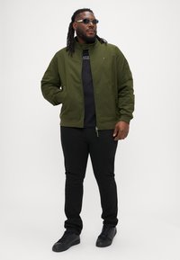 Grøn bomberjakke i let materiale med høj krave, lynlås og elastiske manchetter, parret med en sort skjorte og sorte bukser.