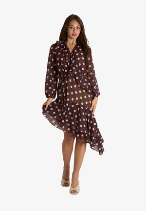 Donna che indossa un vestito a portafoglio marrone con pois bianchi e orlo asimmetrico arricciato, posando con una mano che solleva la gonna.
