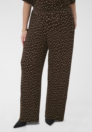 Pantalon marron à jambes larges avec de petits pois beige clair, porté avec des chaussures noires pointues et un bracelet en or au poignet gauche.