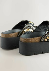 Sandalias de plataforma negras con una plantilla de corcho texturizado y tiras adornadas con cuentas de plata y oro en un patrón ondulado.