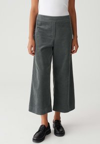 Personne portant un pantalon en velours côtelé gris, large et court, associé à des chaussures noires à lacets et un haut blanc, se tenant devant un fond clair.