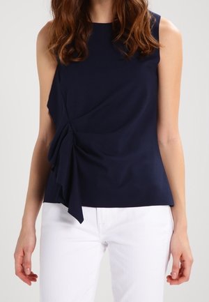 Frau trägt ein ärmelloses, marineblaues Oberteil mit gerafftem Seitendetail, kombiniert mit weißen Hosen, steht vor einem schlichten Hintergrund.