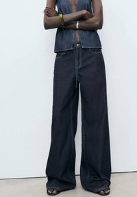 Weit geschnittene dunkle Jeans mit hoher Taille, ausgestattet mit Kontrastnähten und einem lockeren Schnitt. Kombiniert mit einem ärmellosen Denim-Oberteil.