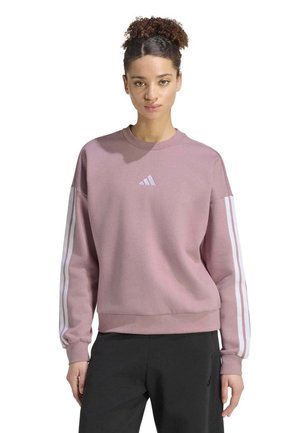 Une femme présente un sweat-shirt Adidas mauve, avec des bandes blanches sur les manches et un pantalon noir, sur fond blanc.