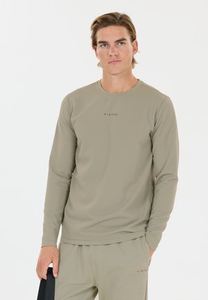 Ung mand iført en matchende beige langærmet skjorte og bukser med "VIRTUS" trykt, står med den ene hånd i lommen og holder en sort genstand.