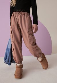 Šviesiai rožiniai kargo kelnių su elastingais manžetais ir keliais kišenėmis, derinami su rudais avies odos šlepetėmis. Modelis laiko džinsinę striukę.