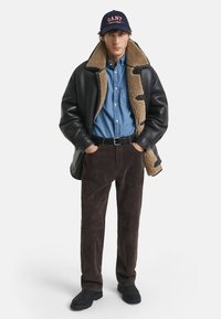 Giubbotto in shearling nero con foderatura beige, camicia in denim blu, pantaloni in suede marrone e stivali neri, completato con un berretto blu navy con il logo "GANT".
