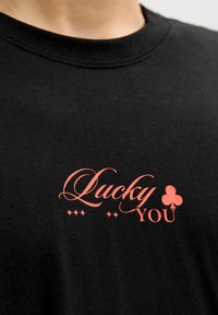 Zwarte katoenen t-shirt met opvallende, koraalkleurige tekst die "Lucky YOU" zegt, vergezeld van een klein klavertje vier en decoratieve stippen.