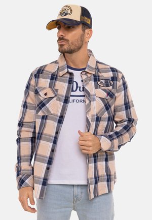 Homme portant une chemise à carreaux beige, bleu marine et grise sur un t-shirt blanc à motif, un jean bleu clair et une casquette de baseball beige et bleu marine.
