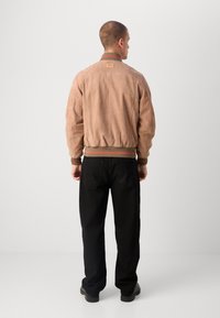 Braune Wildleder-Bomberjacke mit geripptem Kragen und Bündchen, versehen mit einem grün-roten Streifenakzent und einem kleinen Etikett auf der Rückseite. Schwarze Hose sichtbar.