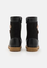 Para czarnych botków do kostki z brązowymi gumowymi podeszwami, zamkiem błyskawicznym oraz małymi tanowymi dekoracjami w kształcie kwiatków po zewnętrznych stronach.