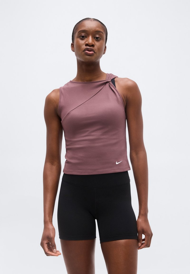 Γυναίκα στέκεται φορώντας αμάνικη μωβ μπλούζα με ασύμμετρο ντεκολτέ και στενά μαύρα σορτς, με φόντο ένα απλό λευκό φόντο.