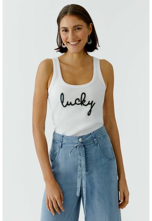 Lächelnde Frau mit kurzen braunen Haaren, die silberne Ohrringe, ein weißes Tanktop mit dem Schriftzug "lucky" und hoch taillierte hellblaue Jeans mit Bindegürtel trägt.