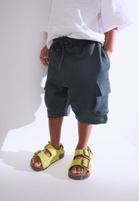 Shorts cargo bleu sarcelle foncé avec une taille élastique, poches latérales, et un t-shirt blanc à manches courtes. Porte des sandales jaunes avec des boucles.