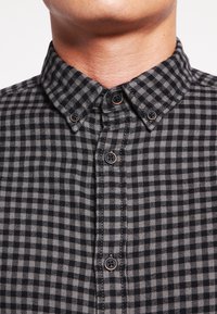 Musta- ja harmaaruudullinen paita, jossa on napillinen kaulus, etunapit ja teksturoitu kangas. Huomiota herättävät tummat napit korostavat mallia.