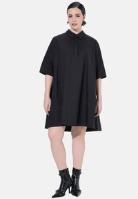 Donna che indossa un vestito nero lungo fino al ginocchio, largo, con maniche a metà e colletto, abbinato a stivaletti neri con tacco alto.