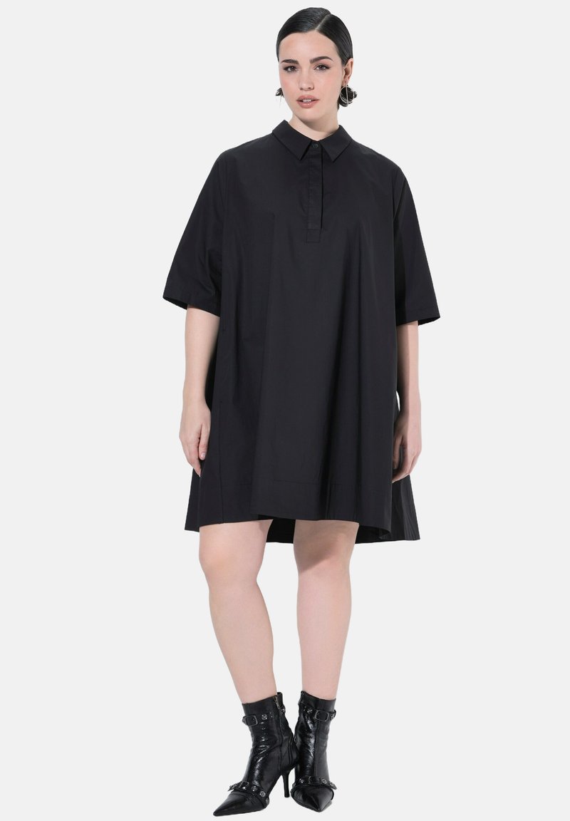 Donna che indossa un vestito nero lungo fino al ginocchio, largo, con maniche a metà e colletto, abbinato a stivaletti neri con tacco alto.