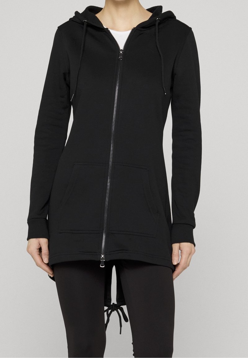 Femme portant un sweat à capuche noir zippé avec cordons et poches avant, associé à un legging noir, sur un fond gris uni.