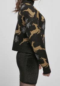 Pull en maille noire avec des motifs dorés de rennes et de flocons de neige blancs. Le pull a un col montant et une coupe décontractée.