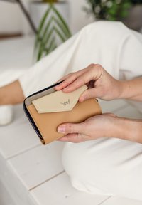 Tan and cream leather wallet with a zip closure, featuring a triangular flap and a small emblem. Held in hands against a light background.