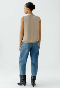 Beige ärmellose Bluse aus leichtem Stoff mit gerafftem Saum; kombiniert mit weit geschnittenen blauen Jeans und schwarzen Ankle-Boots. Schlichte, neutrale Töne.