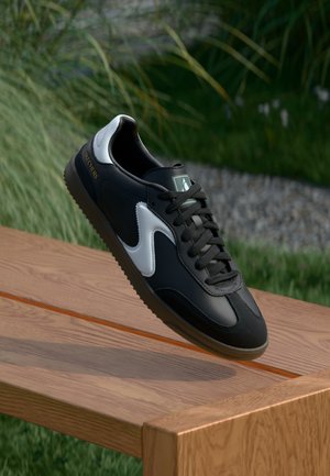 Baskets Skechers noires et blanches avec des lacets reposant sur le coin d’un banc en bois en extérieur avec de l’herbe et des pierres en arrière-plan.