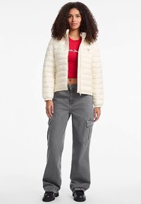 Giacca imbottita color crema con collo alto, abbinata a una camicia corta rossa e pantaloni cargo grigi. Stivali neri massicci completano il look.