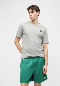 Νέος άνδρας που φοράει γκρι μπλουζάκι Nike και πράσινα σορτς Nike, στέκεται με το ένα χέρι πίσω από την πλάτη του, κοιτάζοντας προς τα αριστερά του.