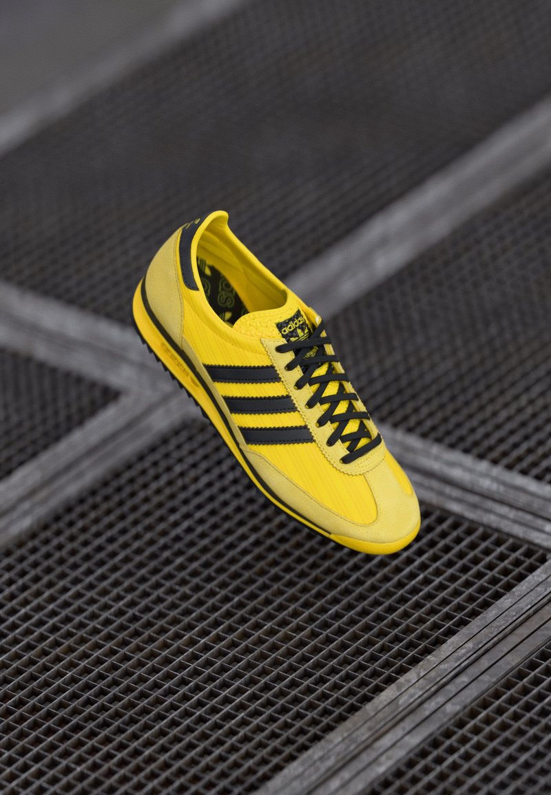 Zapatilla deportiva amarilla con franjas negras, parte superior texturizada, puntera redondeada y cordones negros. Presenta un cuello acolchado y una plantilla con patrón.