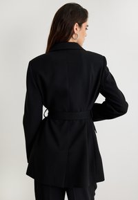 Zwarte op maat gemaakte blazer met een ceintuur in de taille, gestructureerde schouders en een soepele textuur. Heeft een klassieke reverskraag en een middellange stijl.