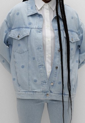 Osoba nosząca jasnoniebieską kurtkę dżinsową z małymi niebieskimi logo, białą koszulę zapinaną na guziki, pasujące jasnoniebieskie dżinsy i długie czarne warkocze zwisające w dół.