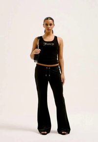 Žena v čiernom tielku s nápisom "Juicy", čiernych širokých nohaviciach, čiernych sandáloch a drží malú čiernu kabelku cez rameno.