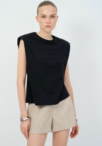 Top nero senza maniche con superficie testurizzata caratterizzata da accenti sferici, abbinato a pantaloni corti beige chiaro. Il tessuto appare liscio e aderente.