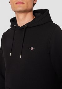 Sudadera negra hecha de tela suave, con un bolsillo frontal, cordones con puntas de metal plateado y un logo bordado en el pecho.