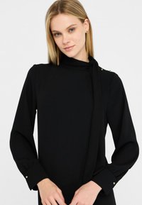 Blusa nera a maniche lunghe con collo alto e dettaglio con fiocco. Presenta accenti in bottone sulle spalle e hardware color oro sui polsi.