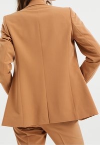Tan tailored blazer with a structured shape, single vent at the back, and smooth texture. Features a notched collar and long sleeves.