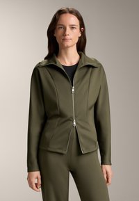 Maslinasto zeleni zip-up jakna s visokim ovratnikom i dugim rukavima, izrađena od glatkog, rastezljivog materijala. Prilagođen kroj, minimalni detalji od metalnih dijelova.
