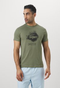 Olive green cotton t-shirt featuring a black Lacoste logo and graphic print at the center. Simple crew neck and short sleeves.