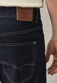 Tamno plave traperice s kontrastnim žutim šavovima, ko�žnom oznakom na struku i klasičnim dizajnom s pet džepova.